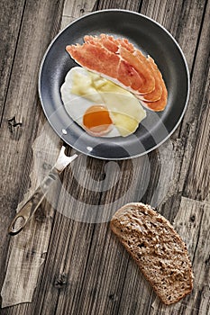 Prosciutto Rashers With Fried Egg In Frying Pan With Slice Of Brown Bread Set On Old Cracked Wooden Garden Table photo