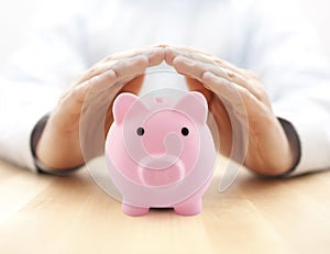 Protect your money. Piggy bank protected by hands on wooden table.