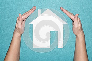 Protection of the house concept. Insurance of real estate. Hands guard white paper house on blue background.