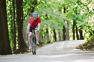 In protective eyewear. Cyclist on a bike is on the asphalt road in the forest at sunny day