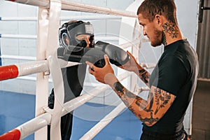 In protective helmet and with glowes. On the ring. Coach is teaching the boy box techniques indoors