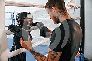 In protective helmet and with glowes. On the ring. Coach is teaching the boy box techniques indoors