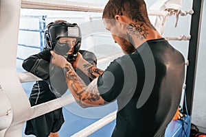 In protective helmet and with glowes. On the ring. Coach is teaching the boy box techniques indoors