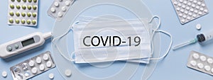 Protective medical face mask, pills, thermometer on blue. Text: COVID-19. Banner.