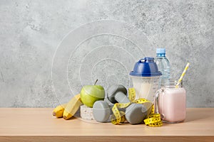 Protein cocktail, dumbbells, measuring tape, water bottle and fresh fruits on wooden table