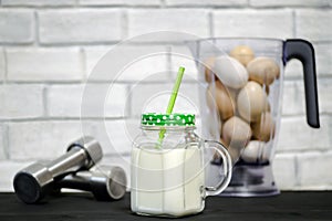 Protein shake in a mug against the background of a blender bowl with chicken eggs and dumbbells against a white wall