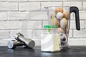 Protein shake in a mug against the background of a blender bowl with chicken eggs and dumbbells against a white wall