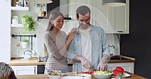 Proud loving young woman hugging watching beloved husband preparing meal.