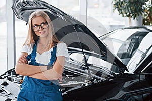 Proud of successful job. Female repairer in the white shirt and blue uniform stands against black car with hood up