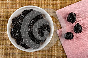 Prune in white bowl, dried plums on paper napkin on mat. Top view