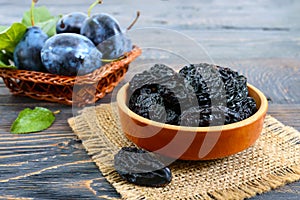 Prunes in a clay bowl and fresh plums, leaves on a wooden table