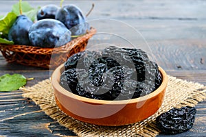 Prunes in a clay bowl and fresh plums, leaves on a wooden table.