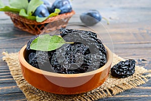 Prunes in a clay bowl and fresh plums, leaves on a wooden table.