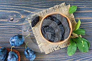 Prunes in a clay bowl and fresh plums, leaves on a wooden table.