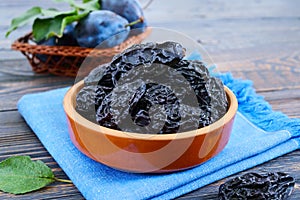 Prunes in a clay bowl and fresh plums, leaves on a wooden table.