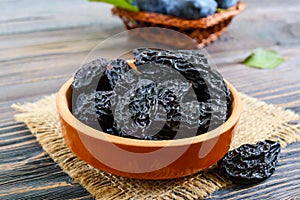 Prunes in a clay bowl and fresh plums, leaves on a wooden table.