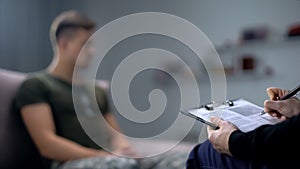 Psychologist making notes during therapy session with sad male soldier, PTSD