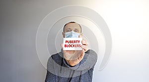 Puberty blockers symbol. White card with words `Puberty blockers`. A young man in a grey wear and medical mask. Sunshine.