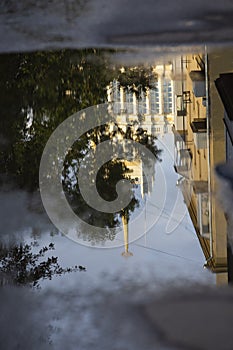 Puddle reflections of walls, windows, roofs of buildings and blue sky on wet asphalt pavement of city street road after summer