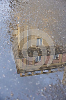 Puddle reflections of walls, windows, roofs of buildings and blue sky on wet asphalt pavement of city street road after summer