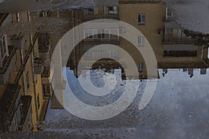 Puddle reflections of walls, windows, roofs of buildings and blue sky on wet asphalt pavement of city street road after summer