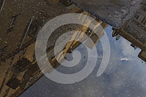 Puddle reflections of walls, windows, roofs of buildings and blue sky on wet asphalt pavement of city street road after summer