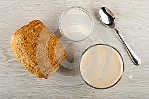 Puff bun, sugar in bowl, spoon, cup with fermented baked milk on wooden table. Top view