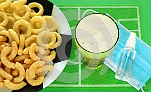 Puffed corn snacks on soccer ball plate, glass of beer, green football field dish mat, blue medical mask and sanitizer