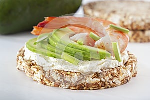 Puffed exploded wheat grains with shrimps and avocado slices on a thin layer of soft cheese on a light wooden table.