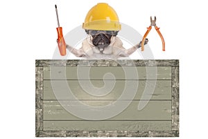 Pug dog holding pliers and screwdriver behind blank old wooden sign, isolated on white background