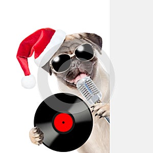 Pug puppy with retro microphone with vinyl record in red santa hat peeking from behind empty board. isolated on white background