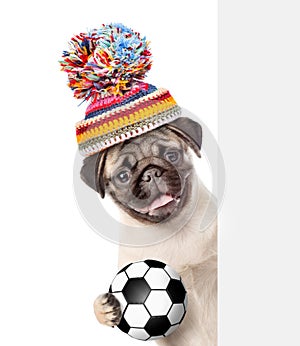 Pug puppy wearing a warm hat with soccer ball peeking from behind empty board. isolated on white background