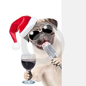 Pug puppy with wineglass and retro microphone in red christmas hat peeking from behind empty board. isolated on white background