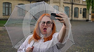 Puggy ginger girl with glasses is making selfie on smartphone in park in rainy weather, fooling around, holding umbrella