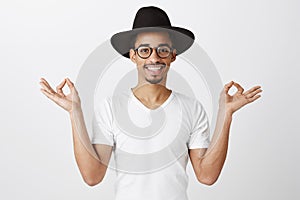 Pulling emotions together. Studio portrait of good-looking african man in trendy eyewear and black hat, raising hands