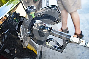 Pumping gas - closeup of gas pump nozzle inserted into to car gas tank with legs of customer in shorts in background