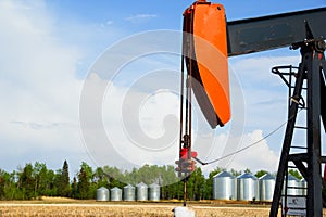 Pumpjack in the agricultural field with steel granaries along the bush
