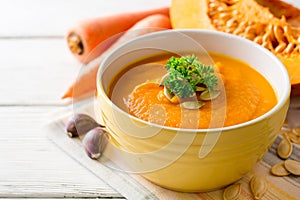 Pumpkin and carrot cream soup with pumpkin seeds and parsley in bowl on white wooden background.