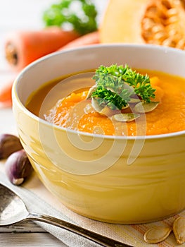 Pumpkin and carrot cream soup with pumpkin seeds and parsley in bowl on white wooden background.