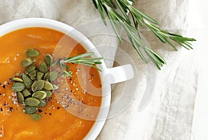 Pumpkin and carrot soup with cream,  seeds and rosemary herb on beige linen background.