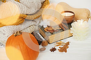 Pumpkin, cozy sweaters, autumn leaves, burning candle and vintage book in golden lights bokeh on white wooden background in room.
