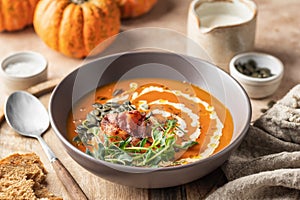 Pumpkin cream soup decorated with cream, pumpkin seeds, bacon, microgreens and olive oil on brown textured background
