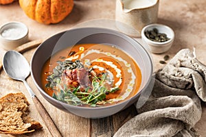 Pumpkin cream soup decorated with cream, pumpkin seeds, bacon, microgreens and olive oil on brown textured background