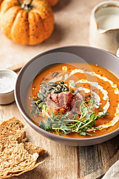 Pumpkin cream soup decorated with cream, pumpkin seeds, bacon, microgreens and olive oil on brown textured background