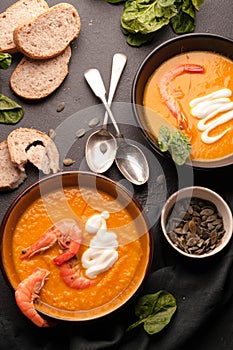 Pumpkin cream soup with shrimps and sour in dark bowls near homemade bread, seeds, herbs