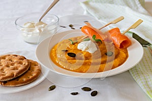 Pumpkin cream soup with whipped cream on white blanket with salmon rolls on the wooden skewer.