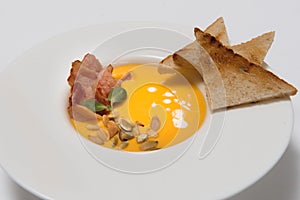 Pumpkin cream soup. White ceramic bowl with fresh pumpkin, dry pumpkin seeds, smoked bacon on a white background
