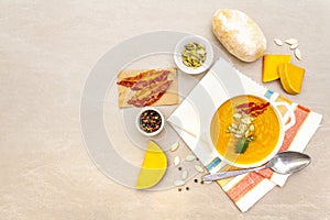 Pumpkin cream soup. White ceramic bowl with fresh pumpkin, dry pumpkin seeds, smoked bacon and bun roll. With vintage linen