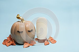 pumpkin with false eyelashes and mock up cosmetic container shampoo or gel on blue background with autumn leaves, natural skin