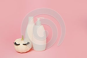 Pumpkin with false eyelashes and mock up cosmetic containers shampoo or gel on pink background, natural skin care in autumn